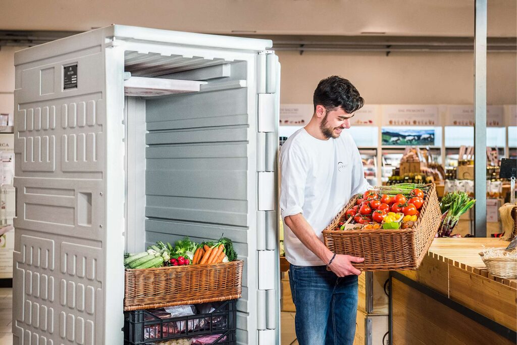 Un homme dispose des légumes devant un meuble réfrigéré professionnel rempli de produits frais.