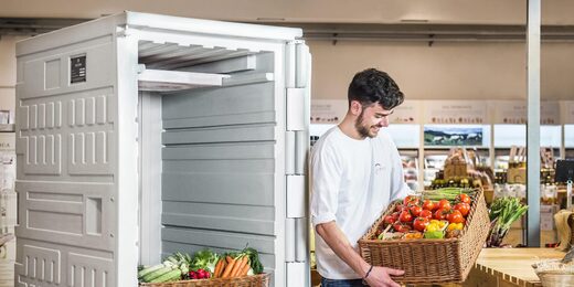 Un homme pose des légumes frais près d’un grand réfrigérateur ouvert dans un magasin.